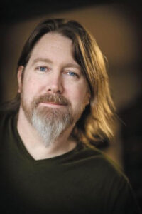 A man with a goatee and long brown hair wearing a black v-neck t-shirt