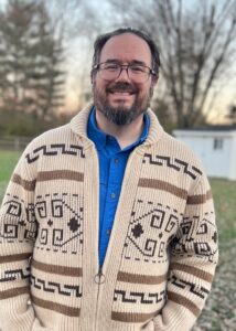 A man in a blue button up and knitted tan and brown sweating standing against a large backyard