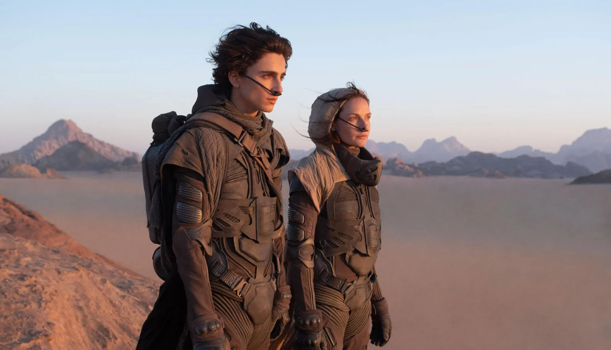 Paul (Timothée Chalamet) and Lady Jessica (Rebecca Ferguson) in sand suits standing on a dune during a sunrise in 'Dune,' 3 Lessons from 'Dune' Screenwriter Eric Roth for Conquering Any Genre (or Planet)