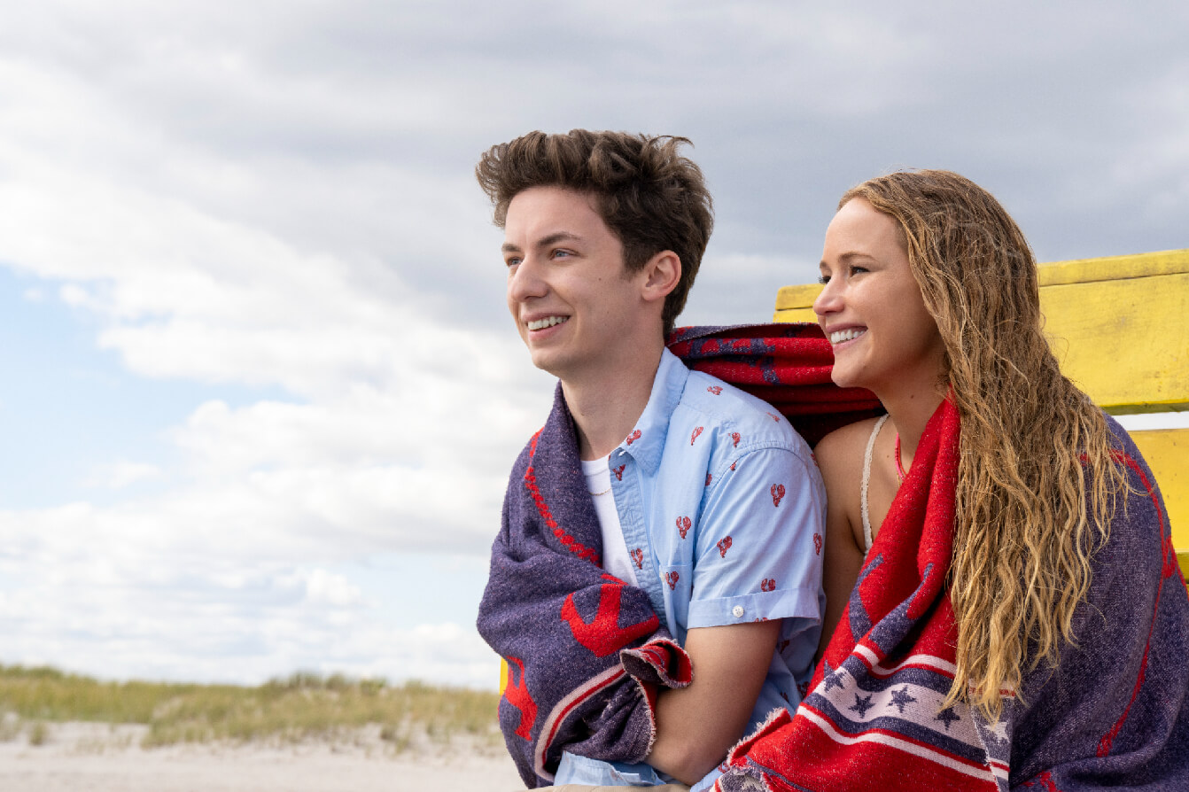 Maddie (Jennifer Lawrence) and Percy (Andrew Barth Feldman) sitting on the beach under a blanket in 'No Hard Feelings,' 5 Insiders to Know if You Write Comedy