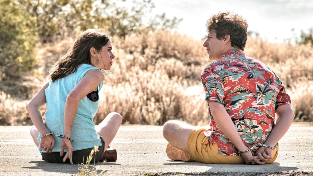 Nyles (Andy Samberg) and Sarah (Cristin Milioti) sitting on the side of the road with handcuffs on in 'Palm Springs,' 100 Meet-Cute Scenarios to Spark Your Screenplay's Romance