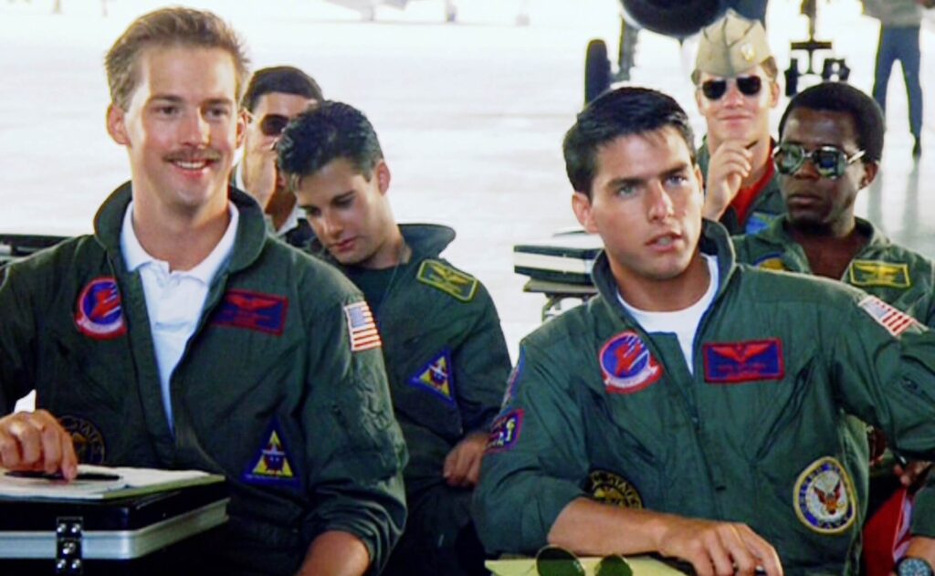 Goose (Anthony Edwards) and Maverick (Tom Cruise) sitting in class in 'Top Gun,' 10 Sidekick Character Archetypes in Movies and TV