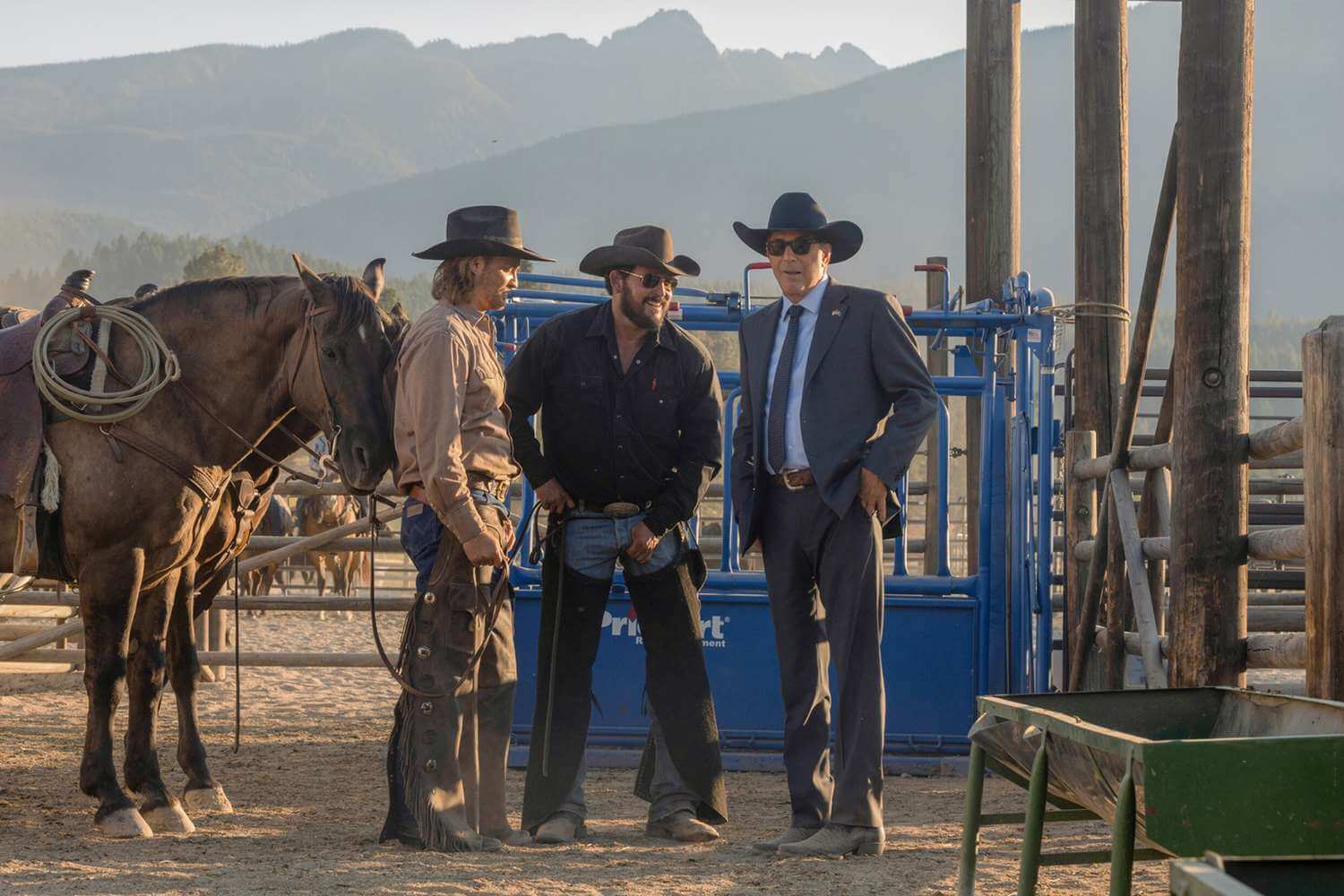 Two cowboys standing next to a man in a black and white suit next to a ranch in 'Yellowstone,' How Taylor Sheridan Redefined the Western Genre With His Own Stories