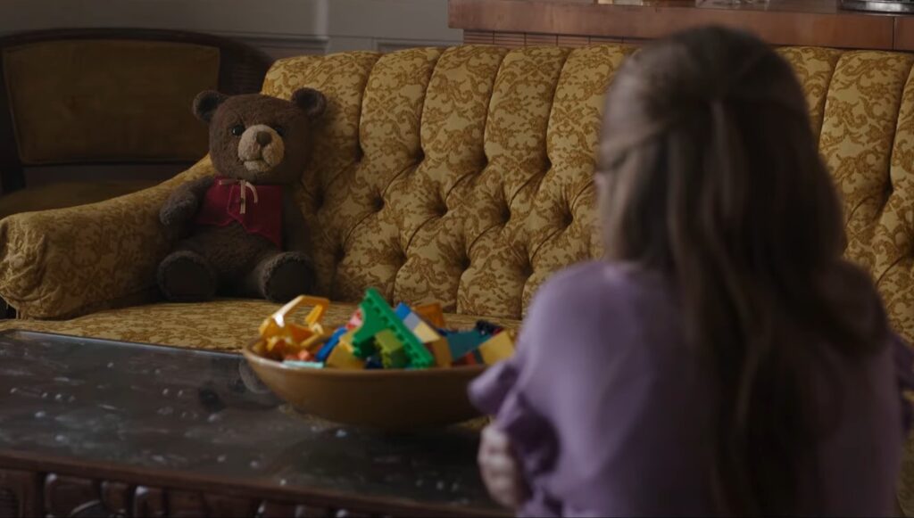 A fearful Alice (Pyper Braun) looking at Chauncey Bear sitting on a yellow couch in 'Imaginary'