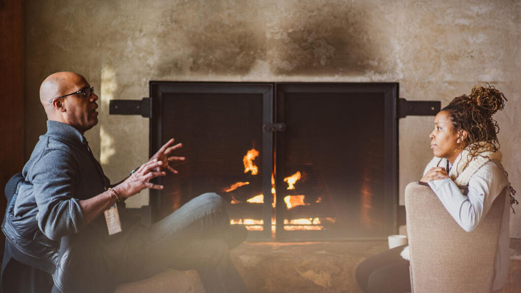 Two people talking in front of a fireplace