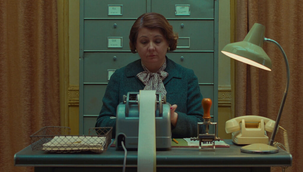 A woman in a blue suit typing behind a desk 'The French Dispatch'