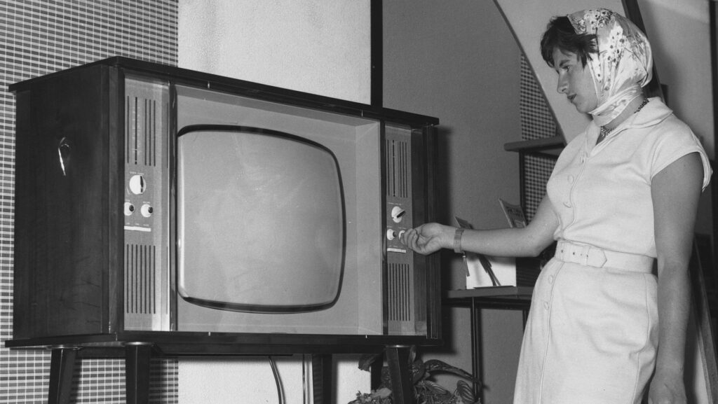 A black and white image of a woman looking at a box TV