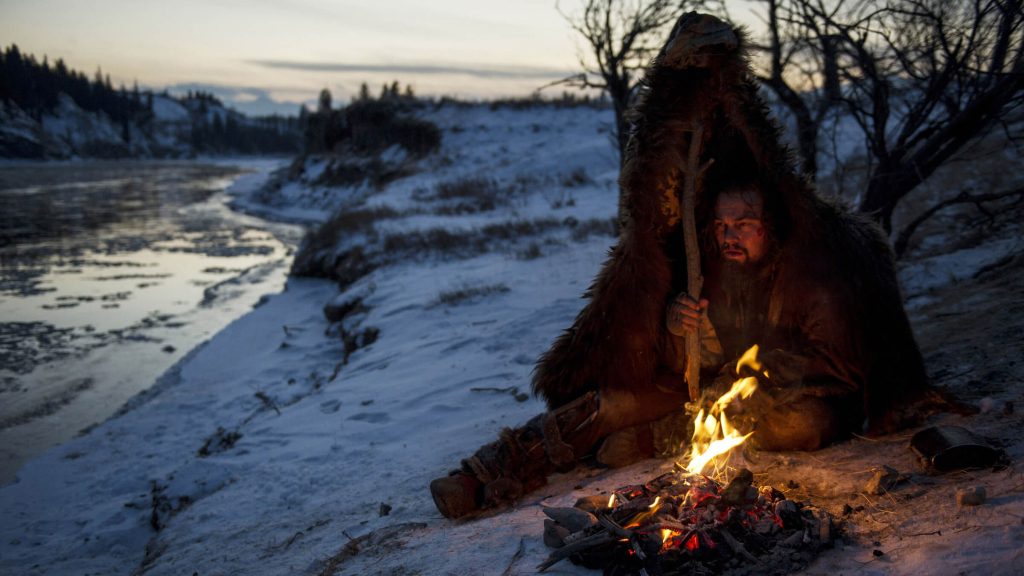 Hugh Glass (Leonardo DiCaprio) sitting by a fire underneath bear fur in 'The Revenant,' 100 Character Vs. Nature Story Prompts