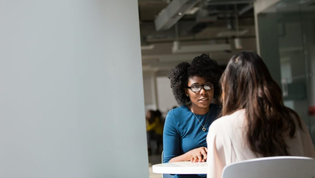 A woman in a blue top with coiled black hair is talking to a woman in a white top with long brown curly hair. 