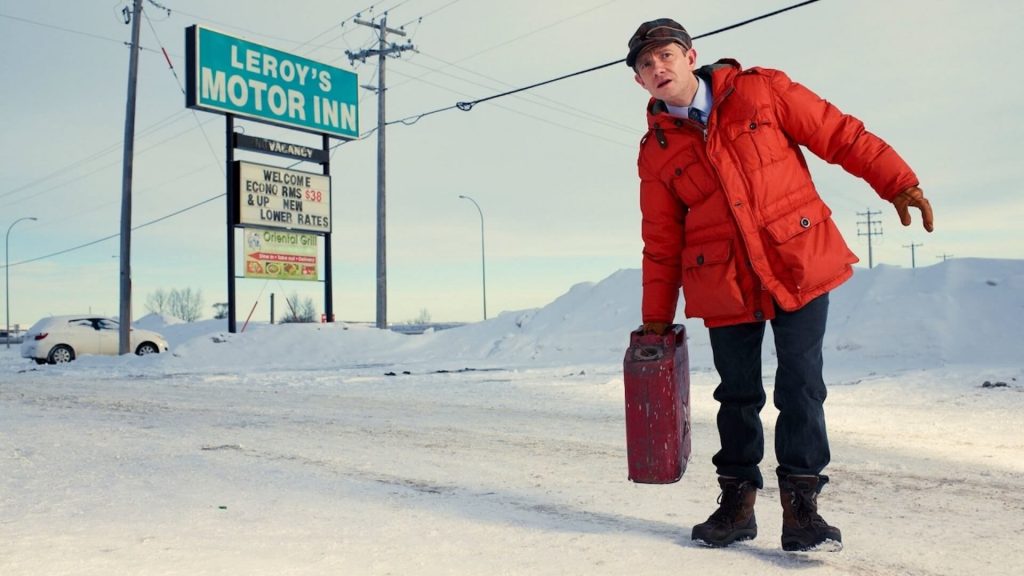 A man in a red jacket with a brief case stands in front of a motel sign in the snow.