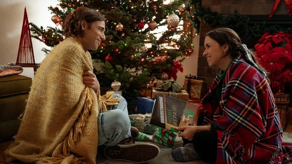 Dustin Milligan as Jake Snowman and Lacey Chabert as Kathy Barrett sit in front of a Christmas tree in 