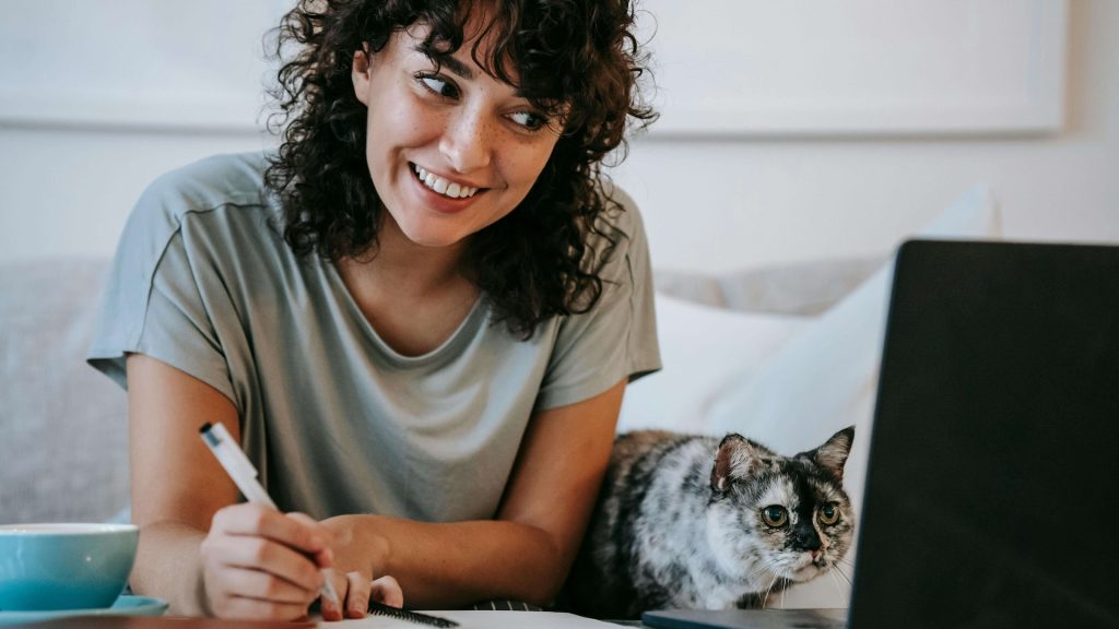 A woman writes notes while looking at a laptop.