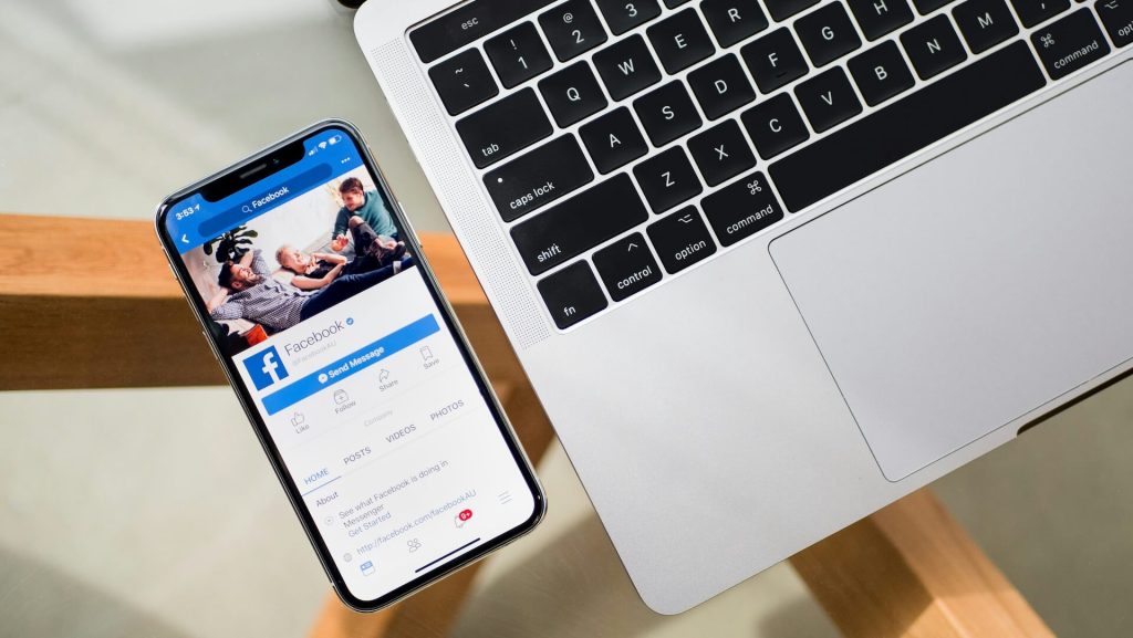 The Facebook app on a smartphone next to a laptop on a glass table.