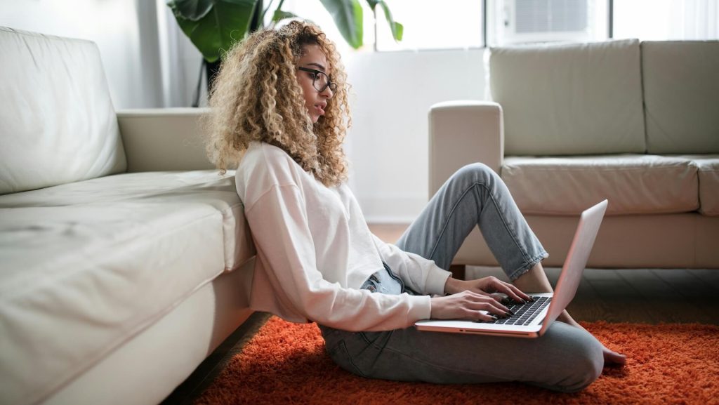 A woman with blonde coiled hair typing on a laptop in a living room.